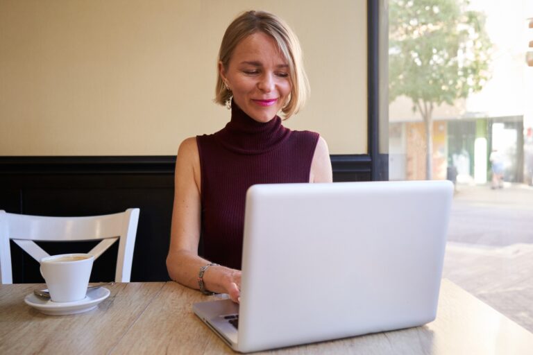 Beautiful mature blonde woman happily telecommuting from coffee shop, using laptop.