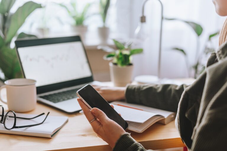 Cropped Freelance woman with mobile phone typing at laptop and working from home office with plants.